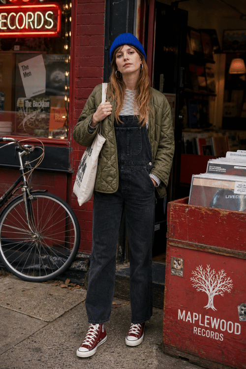 Woman wearing a quilted olive jacket with black denim overalls and red high-top sneakers.