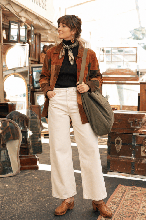 Woman wearing a rust patchwork jacket with white wide leg jeans and brown woven clogs.