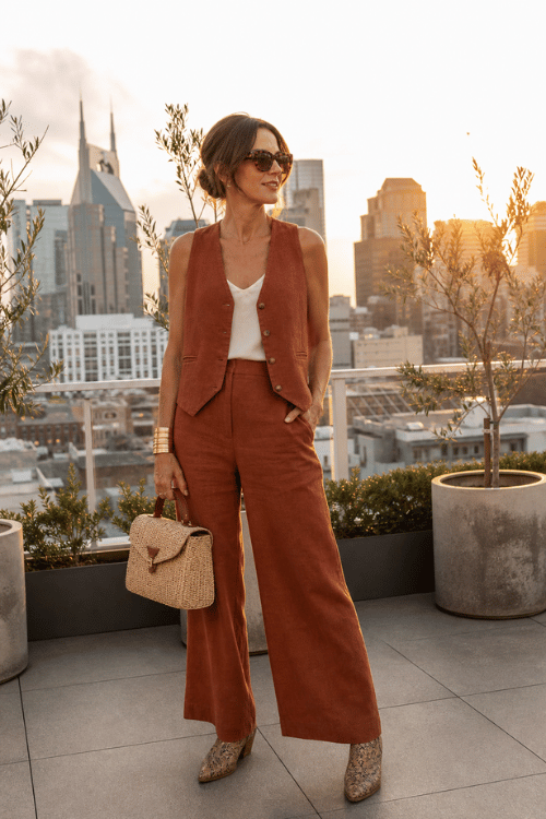 Woman wearing a rust vest with matching wide-leg pants, a white tank top, and snakeskin boots on a rooftop.