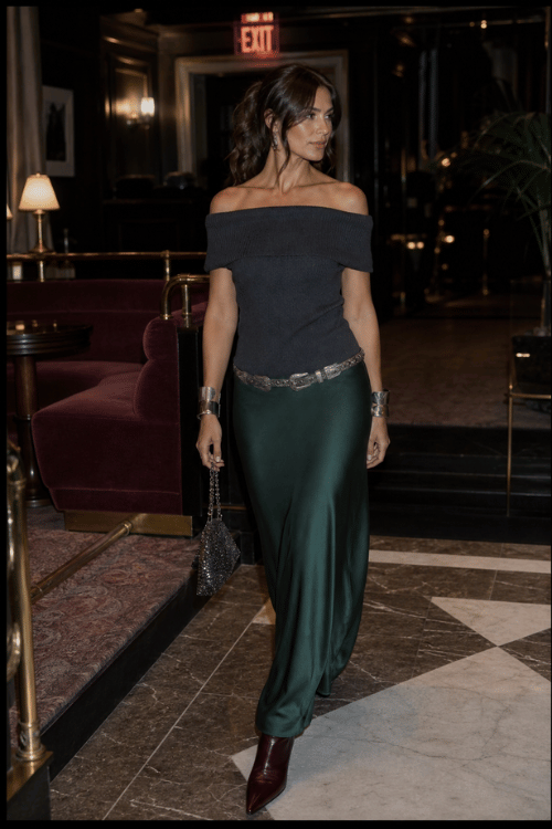 Woman wearing an off-the-shoulder charcoal top with an emerald satin maxi skirt and ankle boots in a luxury hotel.