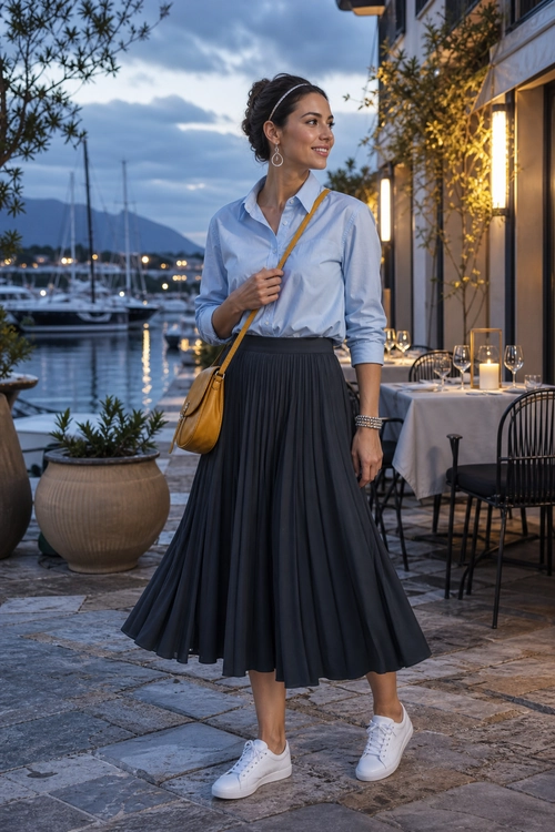 Blue Shirt and Pleated Skirt Outfit Woman wearing a light blue button-up shirt with a dark pleated skirt, white sneakers, and a mustard crossbody bag.