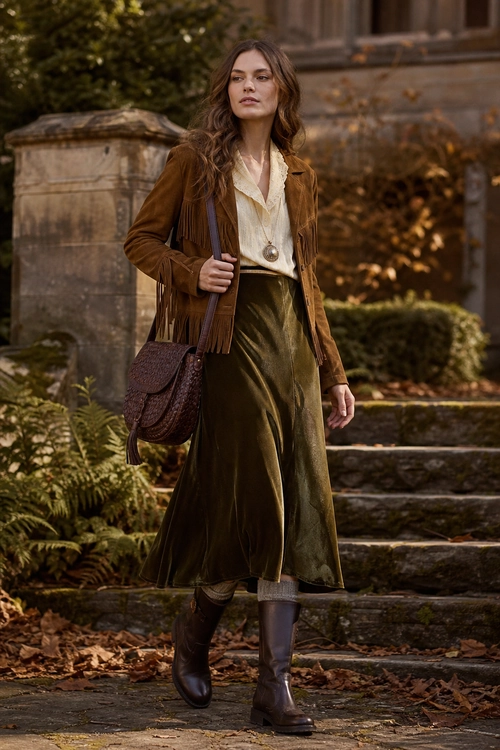 A woman wears a fringed brown jacket over a cream blouse with an olive satin skirt, brown knee-high boots, a pendant necklace, and a shoulder bag.