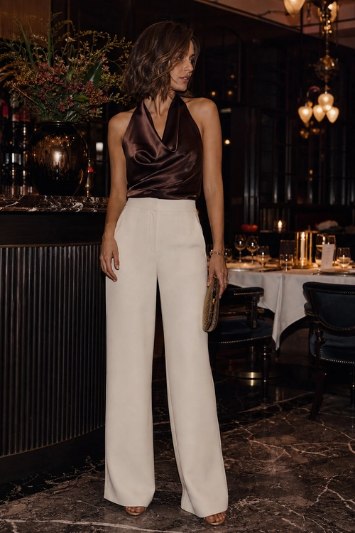 A woman wears a brown satin halter top with ivory wide-leg trousers and carries a small metallic clutch in an elegant restaurant setting.