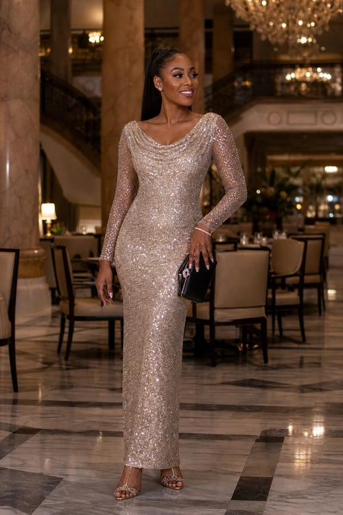 Champagne Sequin Evening Gown A woman wears a fitted champagne sequin gown with long sleeves and strappy heels while carrying a black clutch.