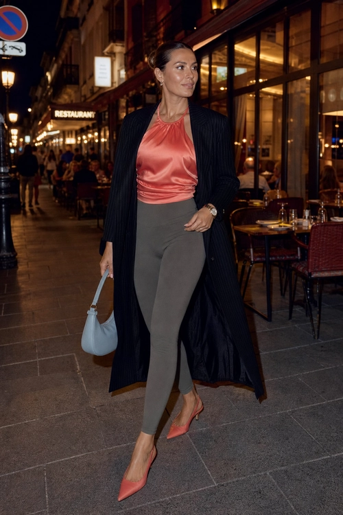 A woman wears a coral satin sleeveless top, long dark pinstripe coat, gray fitted pants, coral pointed heels, and carries a light blue handbag on a city street.