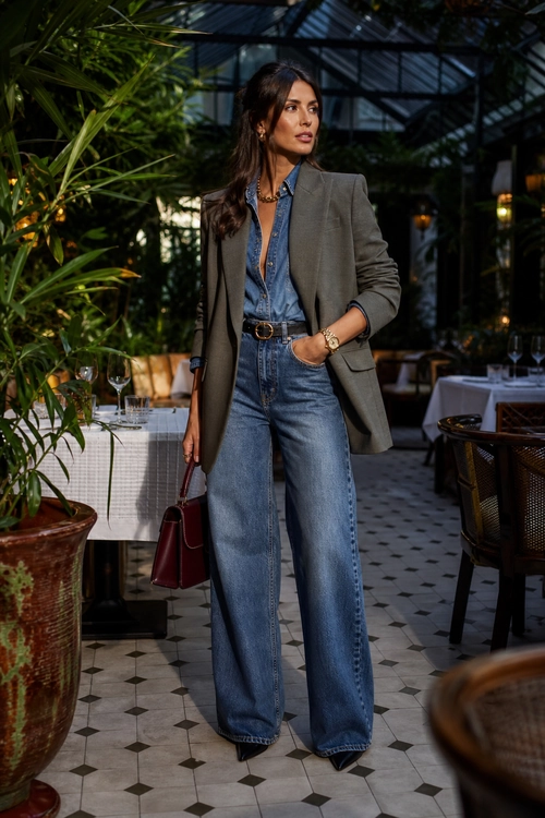 A woman wears a gray blazer over a blue denim shirt with high-waisted wide-leg jeans, a black belt, and a burgundy handbag.