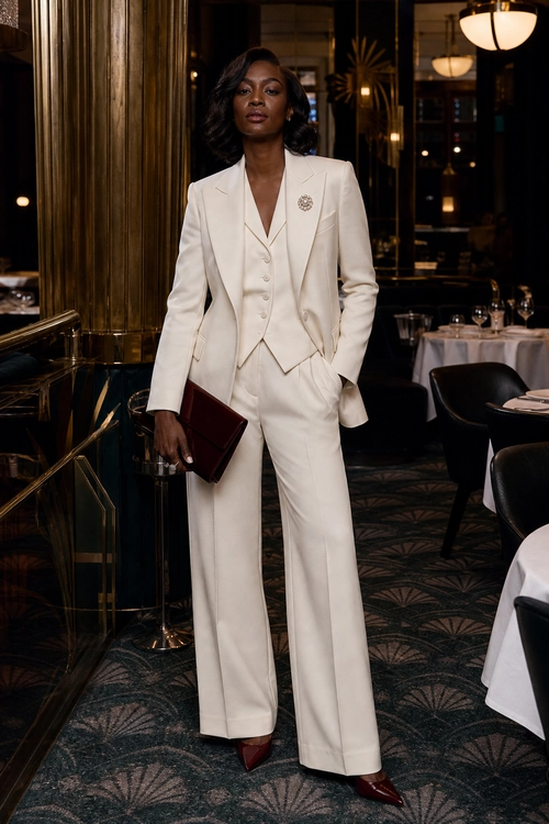A woman wears an ivory tailored blazer, matching waistcoat and wide-leg trousers with burgundy pointed heels and a burgundy clutch.