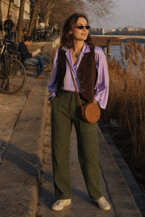Lavender Blouse and Olive Trousers Outfit A woman wears a lavender blouse, brown vest, olive wide-leg trousers, cream sneakers, sunglasses, and a round woven crossbody bag.