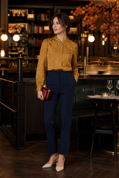 Woman wearing a mustard patterned blouse with navy tailored trousers, cream pointed heels, and a burgundy clutch.