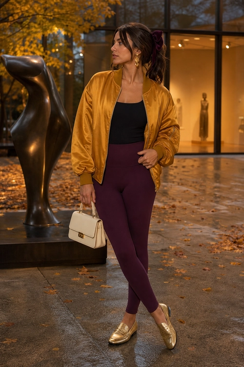 A fashion look featuring a mustard satin bomber jacket, black top, plum leggings, gold loafers, and a cream handbag.