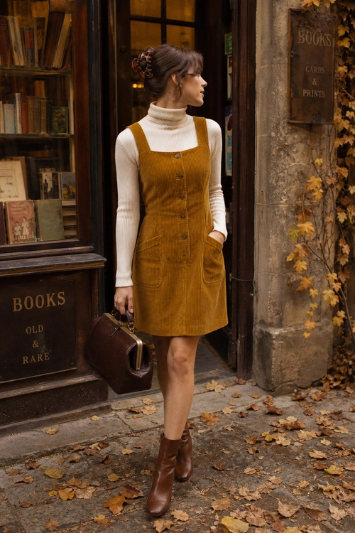 Mustard Corduroy Pinafore Outfit A woman wears a mustard corduroy pinafore dress over a cream ribbed turtleneck with brown ankle boots and a structured brown handbag.