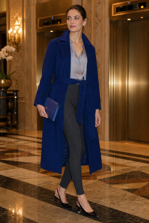 A woman wears a long navy coat over a gray satin blouse and charcoal tailored trousers, styled with black heels and a blue clutch.