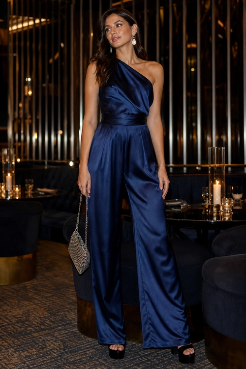 A woman wears a navy satin one-shoulder wide-leg jumpsuit with black platform heels, a silver beaded clutch, and drop earrings.