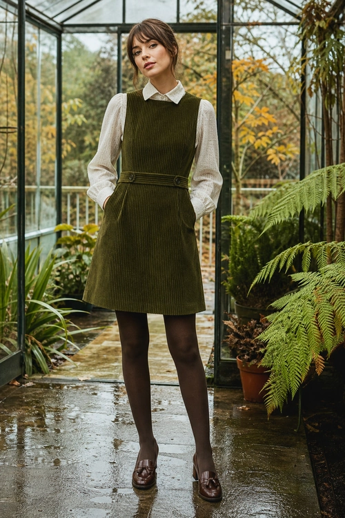 Olive Corduroy Pinafore Outfit A woman wears an olive corduroy pinafore dress over a white collared blouse with dark tights and brown loafers.