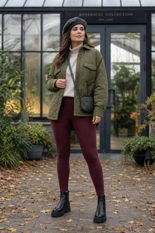 A woman wears an olive quilted jacket over a cream turtleneck with burgundy leggings, a black beret, crossbody bag, and black ankle boots.