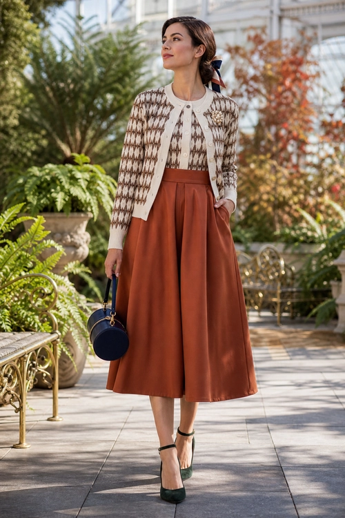 Patterned Cardigan with Rust Midi Skirt A woman wears a cream and brown patterned cardigan with a rust-colored pleated midi skirt, dark heels, and a round navy handbag.