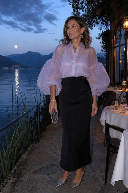 A woman wears a sheer pale pink blouse with voluminous sleeves, a fitted black midi skirt, silver pointed heels, and a transparent clutch.