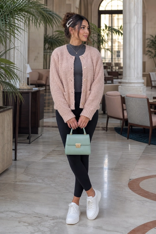 A woman wears a blush pink cardigan over a dark top with black leggings, white sneakers, and a mint green handbag.