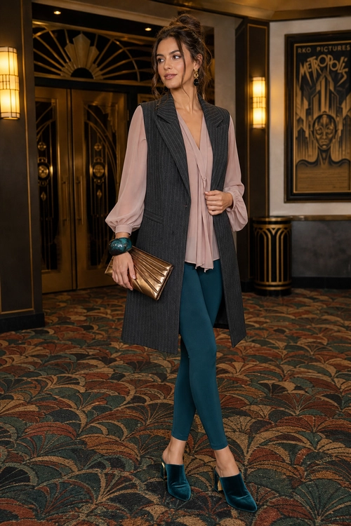 A woman wears a long gray pinstripe vest over a blush pink blouse with teal trousers, teal heels, and a metallic clutch.