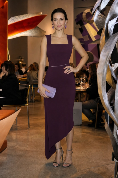 A woman wears a fitted plum sleeveless midi dress with an asymmetrical hem, a lavender clutch, and strappy heels.
