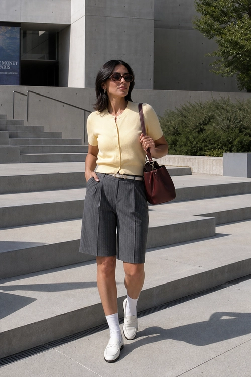 Preppy Bermuda Shorts Outfit A woman wears a pale yellow short-sleeve button top with gray pinstriped Bermuda shorts, white loafers, sunglasses, and a burgundy shoulder bag.