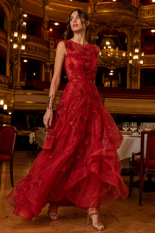 A woman wearing a sleeveless red embellished gown with a flowing sheer skirt, metallic clutch, cuff bracelet, and strappy heels.