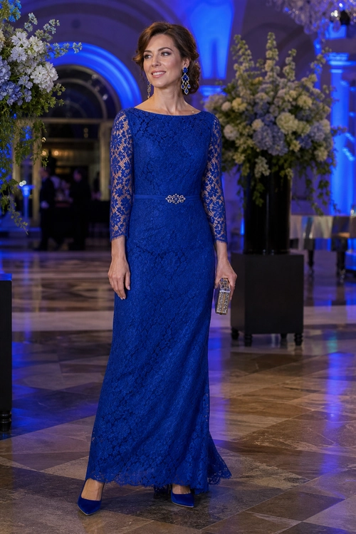 Woman wearing a royal blue floor-length lace gown with sheer three-quarter sleeves, jeweled waist detail, blue heels, drop earrings, and a metallic clutch.