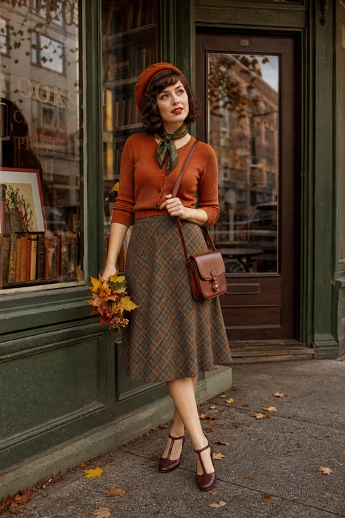 Rust Cardigan and Plaid Skirt Outfit A woman wears a rust cardigan, green plaid midi skirt, neck scarf, beret, brown crossbody bag, and burgundy Mary Jane shoes while holding autumn flowers.