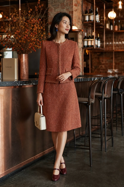 A woman wears a rust textured long-sleeve jacket with a matching knee-length skirt, burgundy Mary Jane heels, and a structured cream handbag.