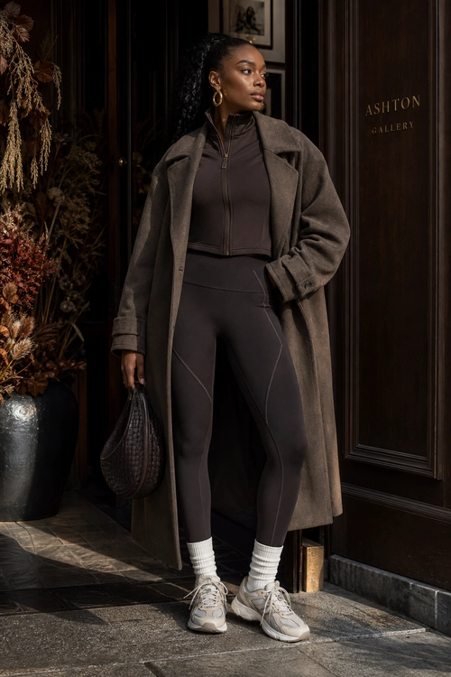 A woman wears a long taupe coat over a dark fitted zip-up top and leggings with white socks, gray sneakers, and a woven handbag.