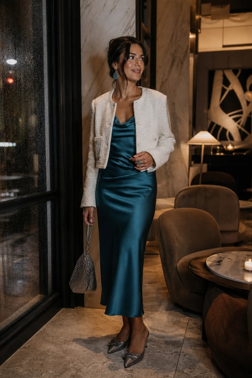 Teal Satin Dress with Cream Jacket A woman wears a teal satin midi dress with a cream textured jacket, pointed shoes, and a small metallic mesh handbag.