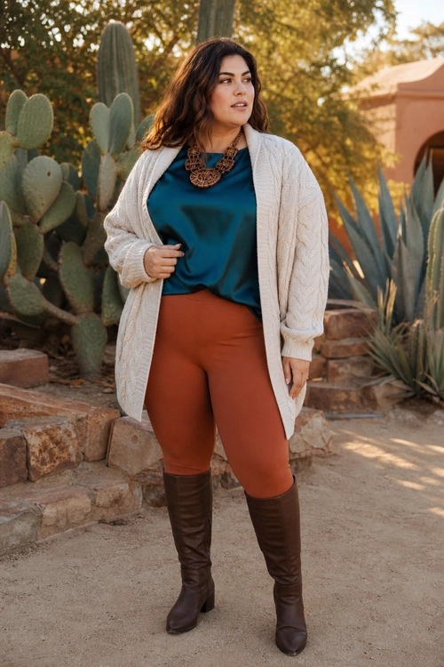 A woman wears a teal top, cream knit cardigan, rust-colored fitted pants, statement necklace, and tall brown boots.