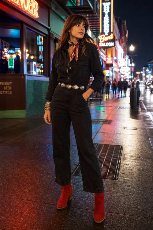 Western-Inspired Black Jumpsuit A woman wears a black button-front denim jumpsuit with a red neck scarf, silver concho belt, stacked bracelets, and red ankle boots on a neon-lit street.
