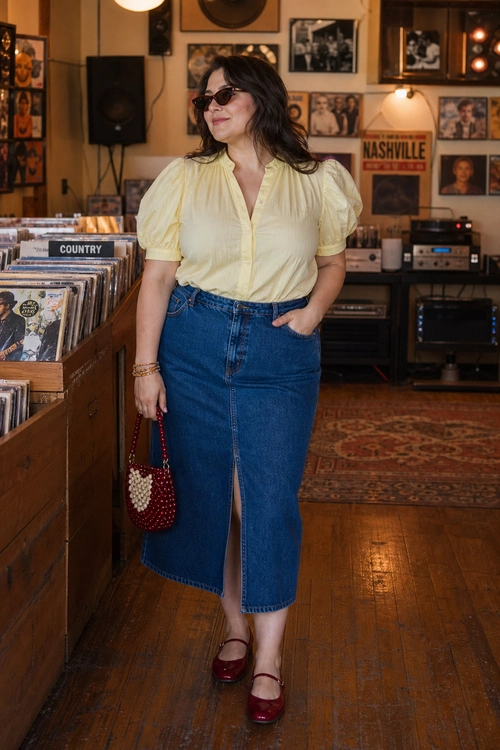 Puff Sleeve Blouse and Denim Midi Skirt A woman wears a pale yellow puff sleeve blouse tucked into a blue denim midi skirt with a front slit, styled with burgundy Mary Jane shoes, sunglasses, and a small red beaded bag.