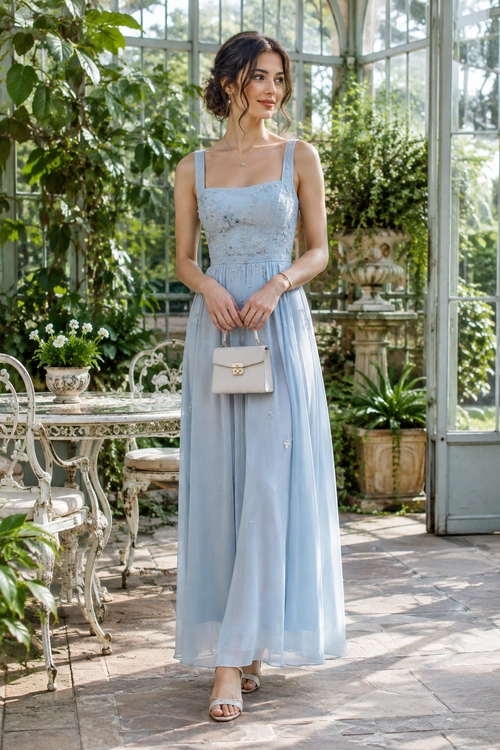 A woman wears a light blue embroidered sleeveless maxi dress with a small beige handbag and neutral sandals.